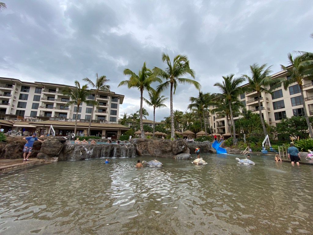 A resort pool area with people swimming and relaxing, surrounded by palm trees and buildings, featuring a small waterfall and a water slide.