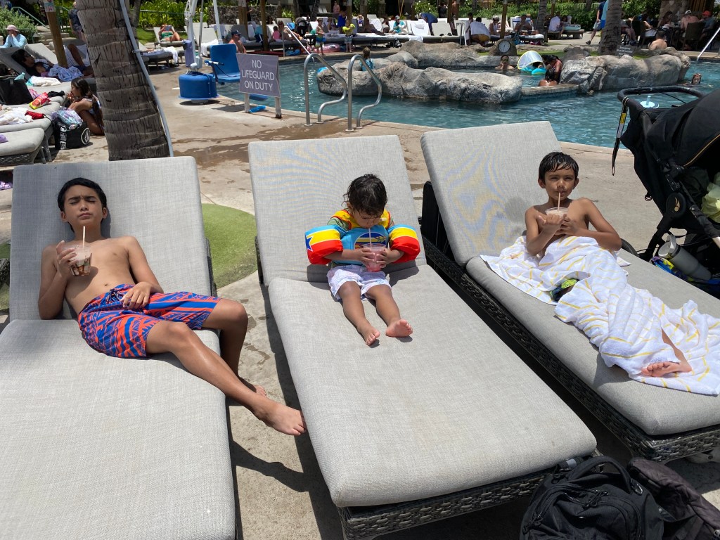 Three children relaxing on lounge chairs by a pool, with one child wearing a colorful floatie and holding a drink.