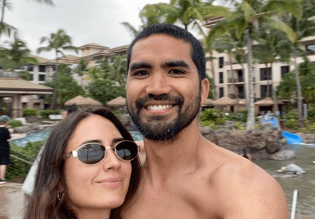 A couple smiling for a selfie in a tropical resort setting with palm trees and a pool in the background.